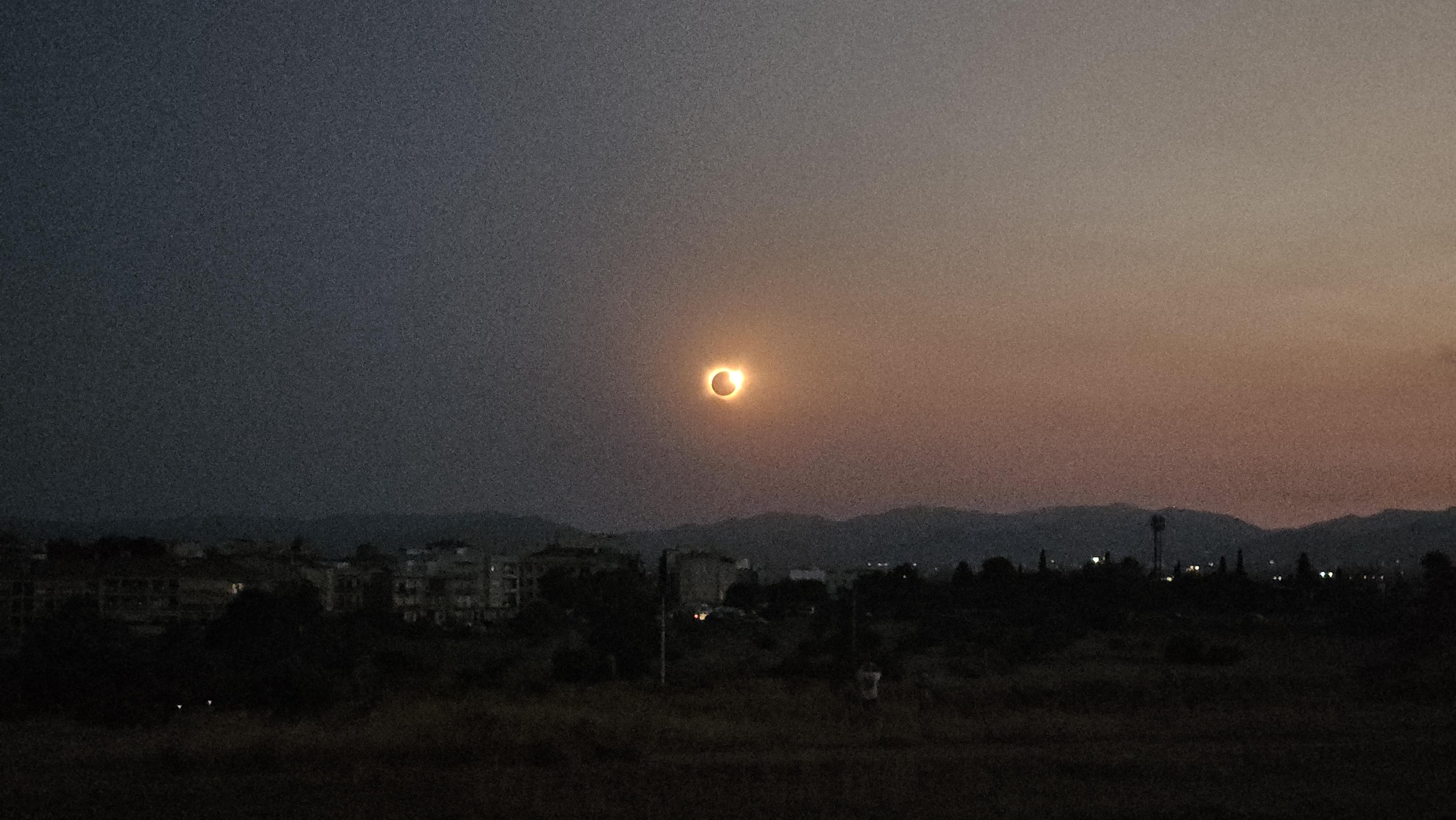 Low-quality phone camera picture showing a ring of fire in the sky as the moon totally covers the sun, with hills, the silhouette of a city and a field seen beneath the sky.